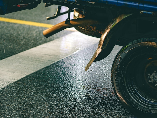 A close-up shot of the rear wheel and exhaust pipe of a vehicle parked on a wet road. The vehicle is positioned on a zebra crossing, and the road surface appears shiny due to rainfall. The tire is labeled with a visible brand name, and the metallic parts show some signs of rust.