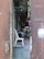 A narrow alley with a tiled floor, lined with clothing hanging on racks. In the background, a person is seated on a white plastic chair, surrounded by various household items. There is another empty chair in the foreground, and the alley appears to be cluttered with everyday objects.