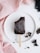 A dark chocolate ice cream bar is placed on a white plate, which is set against a textured pink fabric. Broken pieces of chocolate are scattered around the plate, adding a casual touch to the presentation. In the top corner, a cluster of dark berries is partially visible.