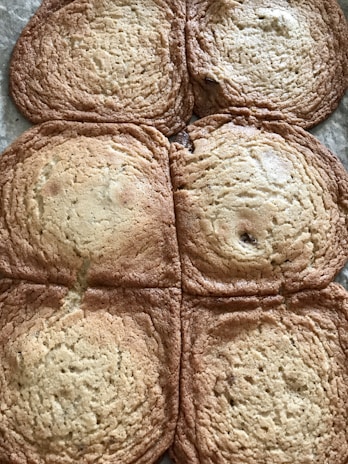 Six large, flat cookies are spread out on a surface. They have a golden-brown color with a slightly darker edge. The cookies appear slightly overbaked or melted together, creating a connected appearance.