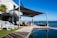 A luxurious outdoor poolside area with modern architecture overlooking the ocean. The scene includes a patio with lounge chairs, a dining table with chairs, and large shade sails providing cover. There are potted plants adding greenery to the setting, and the adjacent infinity pool reflects the clear blue sky.