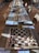 Rows of chess boards are set up on a long table, each equipped with a complete set of chess pieces. The boards are labeled with numbers and situated in a well-lit room. Sunlight casts shadows of the pieces onto the boards and table. Several chairs are arranged around the table.