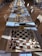 Rows of chess boards are set up on a long table, each equipped with a complete set of chess pieces. The boards are labeled with numbers and situated in a well-lit room. Sunlight casts shadows of the pieces onto the boards and table. Several chairs are arranged around the table.