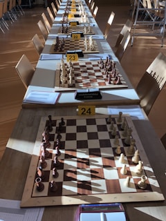 Rows of chess boards are set up on a long table, each equipped with a complete set of chess pieces. The boards are labeled with numbers and situated in a well-lit room. Sunlight casts shadows of the pieces onto the boards and table. Several chairs are arranged around the table.