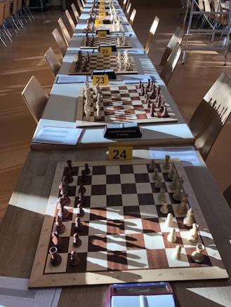 Rows of chess boards are set up on a long table, each equipped with a complete set of chess pieces. The boards are labeled with numbers and situated in a well-lit room. Sunlight casts shadows of the pieces onto the boards and table. Several chairs are arranged around the table.