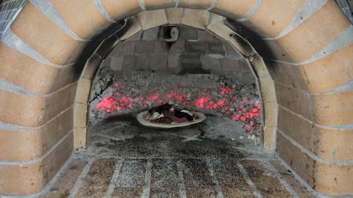 A traditional brick oven with an arched opening contains smoldering red-hot embers. Inside the oven, a pizza on a stone sits close to the embers, absorbing the heat for cooking. The oven is constructed from light brown bricks, and the interior shows signs of use with ash and soot.