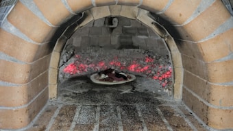 A traditional brick oven with an arched opening contains smoldering red-hot embers. Inside the oven, a pizza on a stone sits close to the embers, absorbing the heat for cooking. The oven is constructed from light brown bricks, and the interior shows signs of use with ash and soot.