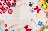 Brightly colored candy canes, red bows, a festive red and white blanket, and several skincare product samples are spread out on a light wooden surface. Christmas-themed items such as a reindeer-patterned blanket and a mug with a reindeer face are included. Small decorations like shiny tinsel, golden stars, and string lights add a festive touch.
