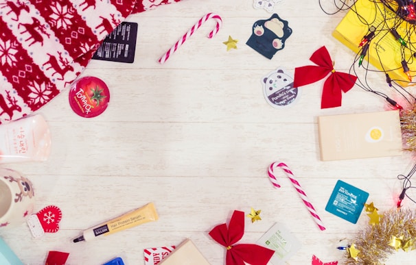 Brightly colored candy canes, red bows, a festive red and white blanket, and several skincare product samples are spread out on a light wooden surface. Christmas-themed items such as a reindeer-patterned blanket and a mug with a reindeer face are included. Small decorations like shiny tinsel, golden stars, and string lights add a festive touch.