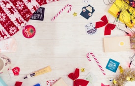 Brightly colored candy canes, red bows, a festive red and white blanket, and several skincare product samples are spread out on a light wooden surface. Christmas-themed items such as a reindeer-patterned blanket and a mug with a reindeer face are included. Small decorations like shiny tinsel, golden stars, and string lights add a festive touch.