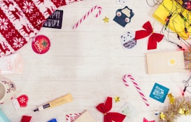Brightly colored candy canes, red bows, a festive red and white blanket, and several skincare product samples are spread out on a light wooden surface. Christmas-themed items such as a reindeer-patterned blanket and a mug with a reindeer face are included. Small decorations like shiny tinsel, golden stars, and string lights add a festive touch.