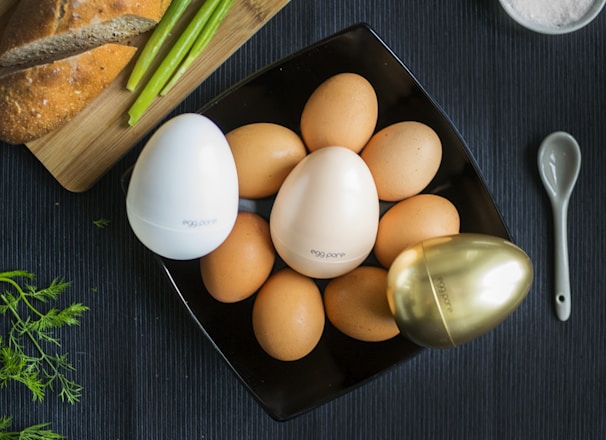 A black square plate holds several brown eggs surrounded by three distinctively designed egg-shaped containers in white, cream, and gold colors, each labeled 'egg pore.' To the side, there is a wooden board with a loaf of bread and some green asparagus. A small, sleek spoon rests nearby on the dark textured surface, and a few sprigs of herbs add freshness.