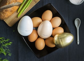 A black square plate holds several brown eggs surrounded by three distinctively designed egg-shaped containers in white, cream, and gold colors, each labeled 'egg pore.' To the side, there is a wooden board with a loaf of bread and some green asparagus. A small, sleek spoon rests nearby on the dark textured surface, and a few sprigs of herbs add freshness.