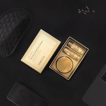 A box of skincare products, specifically labeled as 'Ferment Snail Eye Cream', is open to reveal two small bottles and a compact container. The packaging is golden with a pattern reminiscent of waves or spirals. Surrounding the box, there is a black quilted pouch, a smartphone, black pencils, and part of a laptop, all placed on a black surface.