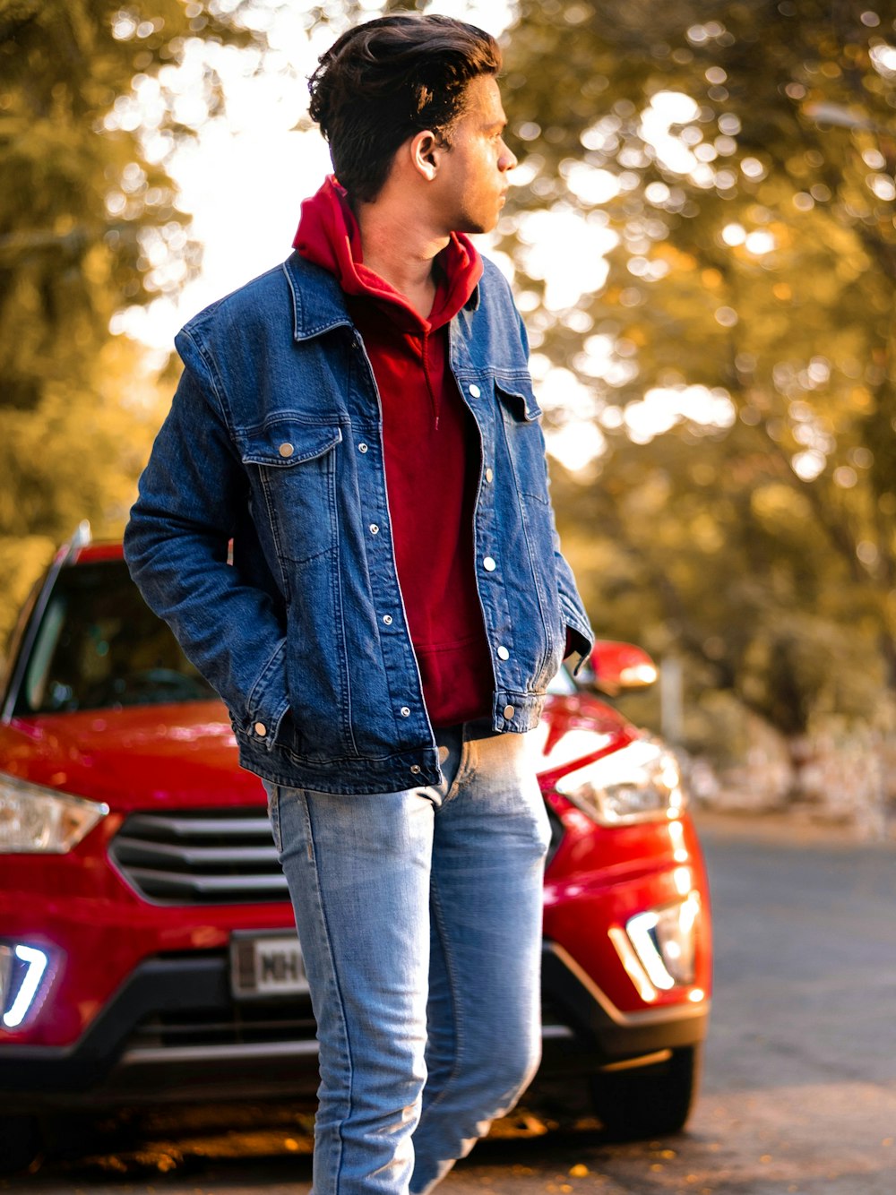 man wearing blue denim jacket standing near red vehicle beside road during daytime