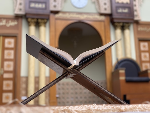 An open book rests on a wooden stand in a setting that appears to be a mosque or prayer area. The background features ornately decorated columns and an arched entrance, suggesting a place of worship. The focus is on the book, which is positioned in the center of the composition, and the intricate details of the surrounding architecture contribute to a serene and respectful atmosphere.