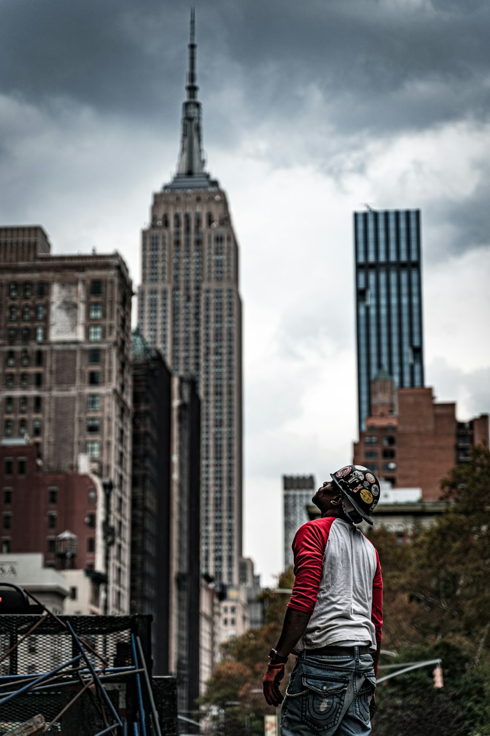 man wearing white and red crew-neck long-sleeved shirt standing and looking up near New York City buildings during daytime