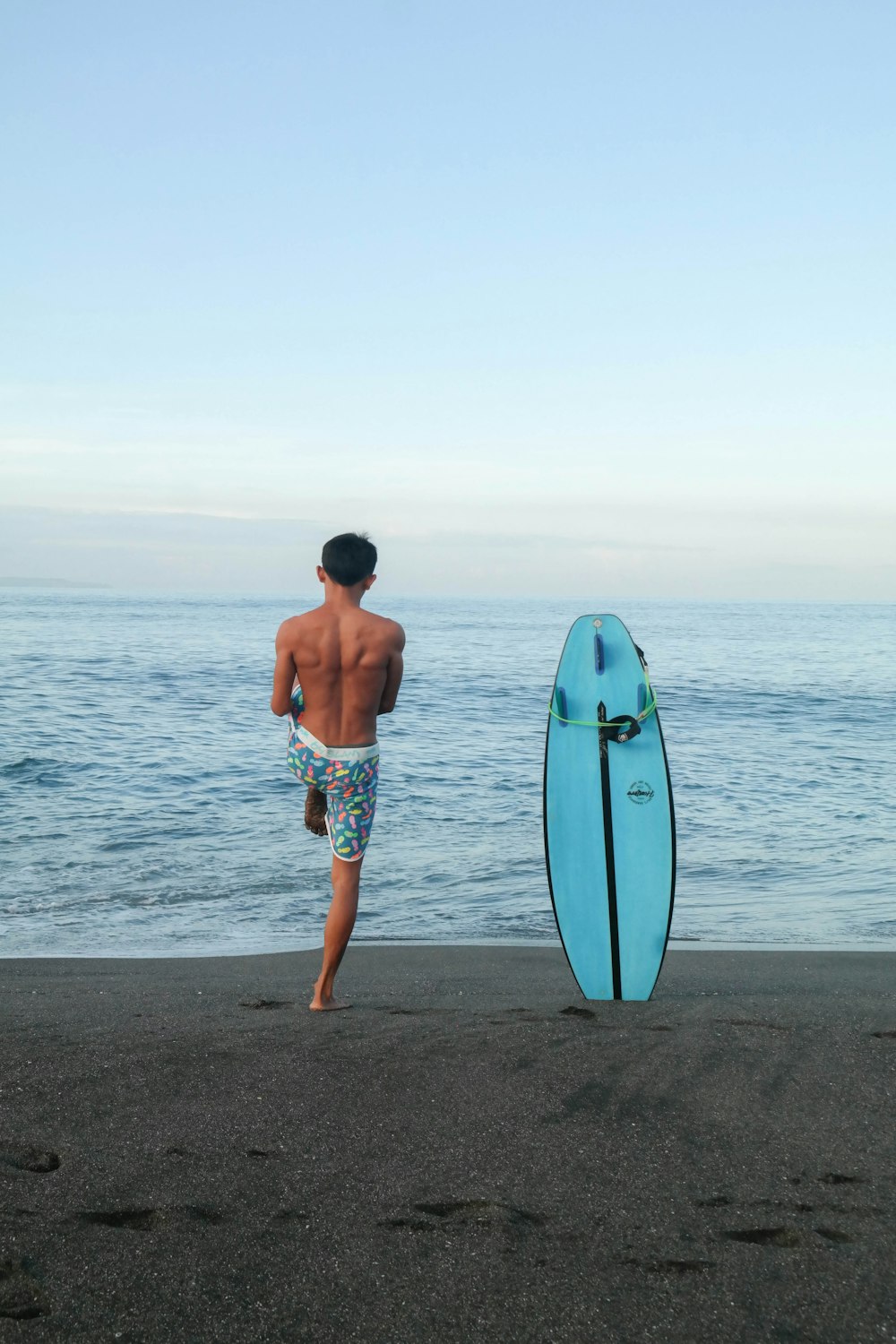 topless man standing near blue surfboard on seashore
