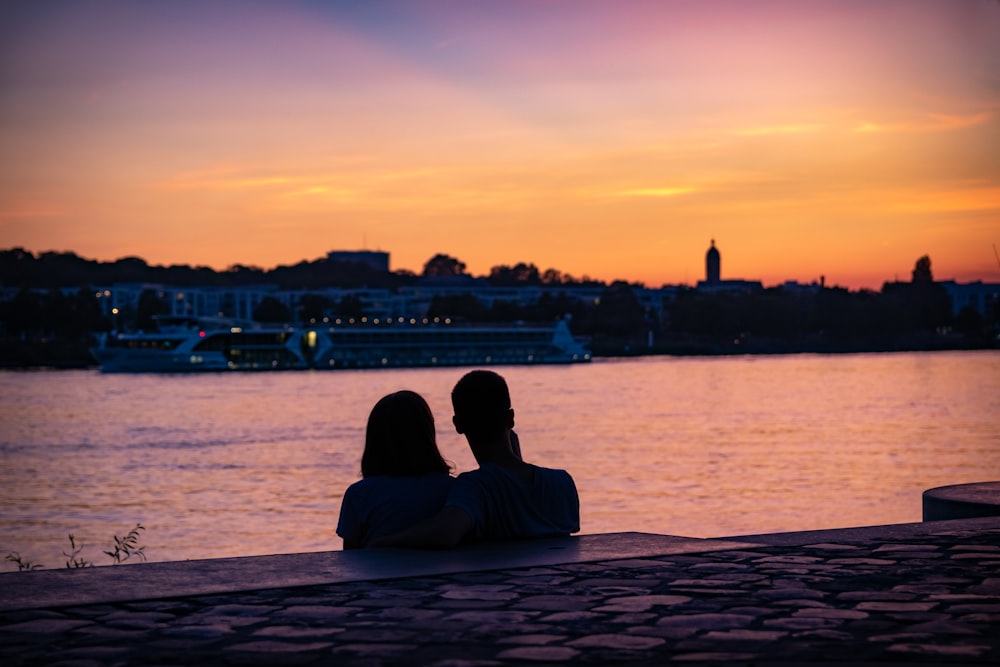 silhouette of couple sitting on shore looking at city skyline during sunset