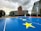 A prominent EU building stands in the background with large glass and steel architecture. In the foreground is a blue sports court adorned with yellow stars, resembling those on the EU flag. The sky is overcast, adding a dramatic effect to the scene. Tall modern office buildings flank both sides of the central structure.