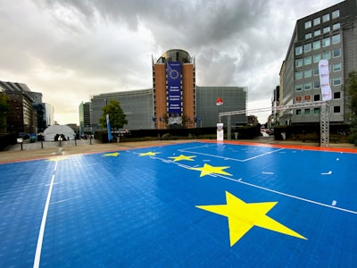 A prominent EU building stands in the background with large glass and steel architecture. In the foreground is a blue sports court adorned with yellow stars, resembling those on the EU flag. The sky is overcast, adding a dramatic effect to the scene. Tall modern office buildings flank both sides of the central structure.