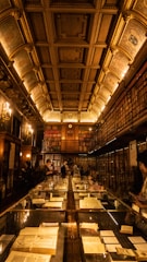 An ornate and historic library with a high, decorative ceiling illuminated by warm lighting. Shelves of books line both sides of the room, and glass display cases filled with antique books and manuscripts run down the center. People are quietly observing the exhibits.