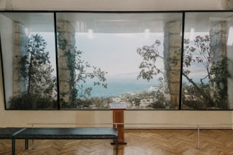 A large painting or window displays a scenic view of the sea framed by stone columns and tree branches. The interior space has a wooden floor and a bench facing the view, suggesting a gallery or museum setting.