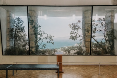 A large painting or window displays a scenic view of the sea framed by stone columns and tree branches. The interior space has a wooden floor and a bench facing the view, suggesting a gallery or museum setting.