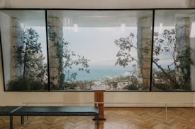 A large painting or window displays a scenic view of the sea framed by stone columns and tree branches. The interior space has a wooden floor and a bench facing the view, suggesting a gallery or museum setting.
