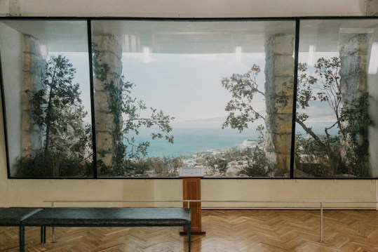 A large painting or window displays a scenic view of the sea framed by stone columns and tree branches. The interior space has a wooden floor and a bench facing the view, suggesting a gallery or museum setting.