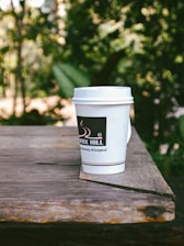 A paper coffee cup with a lid is placed on a weathered wooden surface, set against a backdrop of lush, blurred greenery. The cup bears the logo 'Coffee Hill' and is predominantly white with black accents.