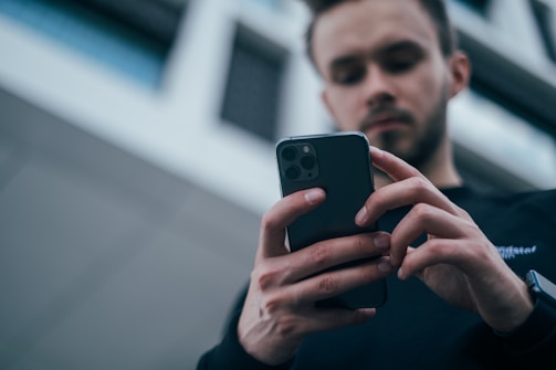 A person is intently using a smartphone, holding it with both hands. The background features a modern building with a slightly blurred facade, suggesting an urban setting. The focus is on the device and the user's engagement with it.