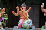 A small, colorful statue depicts a child-like figure with multiple arms sitting on a blue and white striped ball. The figure has an elephant-like face adorned with jewelry, pink clothing, and a traditional headdress. The surrounding area includes other partial figures and some greenery in the background.