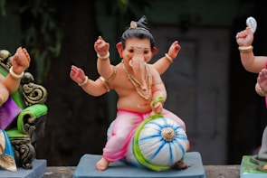 A small, colorful statue depicts a child-like figure with multiple arms sitting on a blue and white striped ball. The figure has an elephant-like face adorned with jewelry, pink clothing, and a traditional headdress. The surrounding area includes other partial figures and some greenery in the background.
