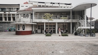 A modern, two-story building with curved and angular design elements, featuring open-air seating areas and industrial aesthetics. The lower level has wide glass doors, and there are several people seated or standing around, engaged in conversation. The upper level is accessible via a white spiral staircase. A few small trees in planters add a touch of greenery to the concrete surroundings.