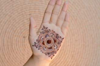 A hand is displayed with intricate henna artwork applied to the palm. The design is symmetrical and features floral and circular motifs in a dark brown color. The hand is positioned against a textured, circular backdrop.