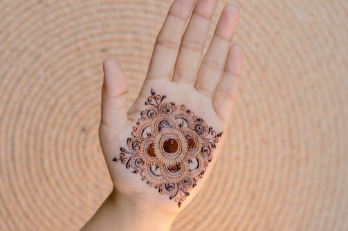 A hand is displayed with intricate henna artwork applied to the palm. The design is symmetrical and features floral and circular motifs in a dark brown color. The hand is positioned against a textured, circular backdrop.