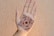 A hand is displayed with intricate henna artwork applied to the palm. The design is symmetrical and features floral and circular motifs in a dark brown color. The hand is positioned against a textured, circular backdrop.