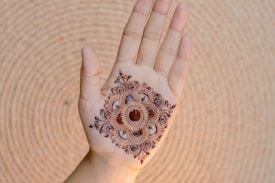 A hand is displayed with intricate henna artwork applied to the palm. The design is symmetrical and features floral and circular motifs in a dark brown color. The hand is positioned against a textured, circular backdrop.