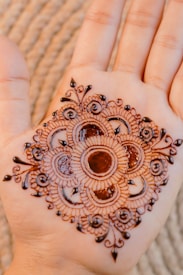 A beautifully intricate henna design is applied to the palm of a hand. The design consists of concentric circles with detailed paisley and floral patterns, featuring delicate lines and dots.