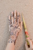 A hand with intricate henna patterns drawn on it, lying against a textured circular mat. The design includes floral motifs and delicate swirls. Beside the hand is a henna cone held by another hand.