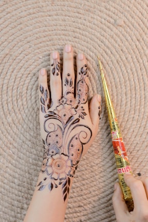 A hand with intricate henna patterns drawn on it, lying against a textured circular mat. The design includes floral motifs and delicate swirls. Beside the hand is a henna cone held by another hand.