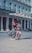 A couple dances energetically in front of a grand hotel, their clothing and movement suggesting joy and connection. The woman's skirt is flowing, and both are smiling, with a backdrop of flags and an elegant building facade.