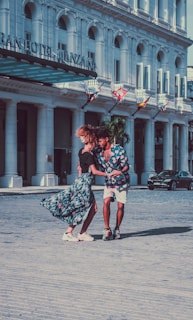 A couple dances energetically in front of a grand hotel, their clothing and movement suggesting joy and connection. The woman's skirt is flowing, and both are smiling, with a backdrop of flags and an elegant building facade.