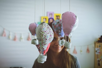 Several handmade fabric hot air balloons hang from strings in the foreground. Each balloon is made from different patterned fabric, including floral, polka dots, stars, and stripes. A person stands in the background with a gentle smile, creating a whimsical and cozy atmosphere. The room is softly decorated, with blurred background elements such as garlands and hanging pictures.