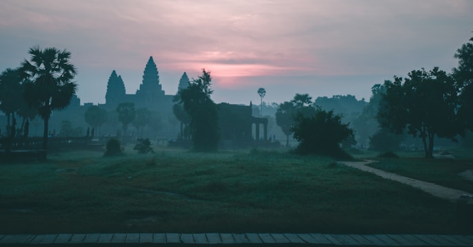 Silhouetted temple structures are set against a misty dawn sky, with soft pink and purple hues from the sunrise. Lush greenery and palm trees surround the area, creating a serene and tranquil atmosphere.
