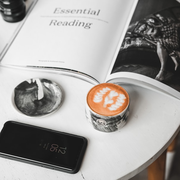A round white table holds a few items including an open magazine, a small black plate, a cup of coffee with latte art, and a smartphone displaying the time as 12:05. The magazine features an article titled 'Essential Reading' with a grayscale photograph of a person. The latte art is shaped like delicate leaves or flowers.