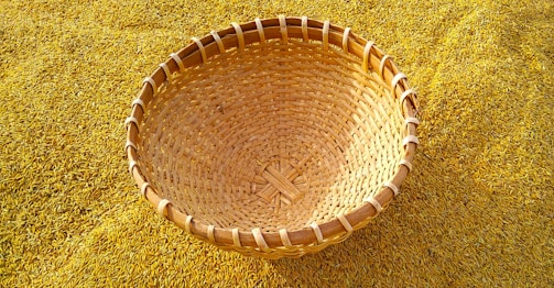 A woven basket is resting on a bed of golden rice grains. The basket is made from natural material and showcases an intricate pattern of weaving. The rice grains are spread densely, creating a textured background that complements the basket's earthy tones.