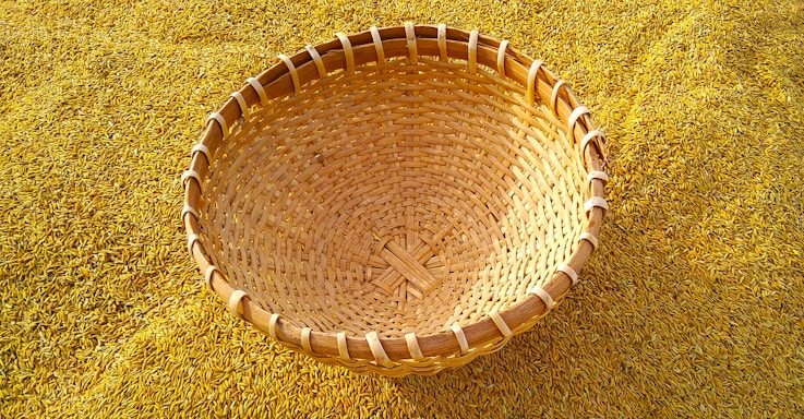 A woven basket is resting on a bed of golden rice grains. The basket is made from natural material and showcases an intricate pattern of weaving. The rice grains are spread densely, creating a textured background that complements the basket's earthy tones.