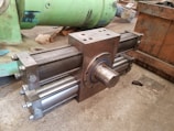 A large, industrial cylinder with multiple metallic rods and bolts is positioned on a concrete floor. The machinery appears robust and heavy-duty, likely used for mechanical or hydraulic purposes. Green and wooden structures are in the background, suggesting a workshop or manufacturing setting.